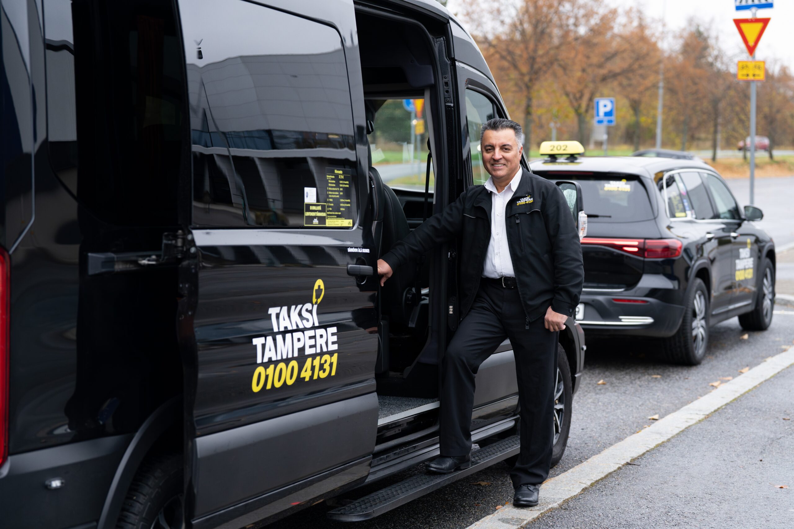 Miestaksinkuljettaja katsoo kameraan ja seisoo iloisesti mustan Taksi Tampere logolla varustetun invataksin vieressä. Taksin sivuovi on auki.